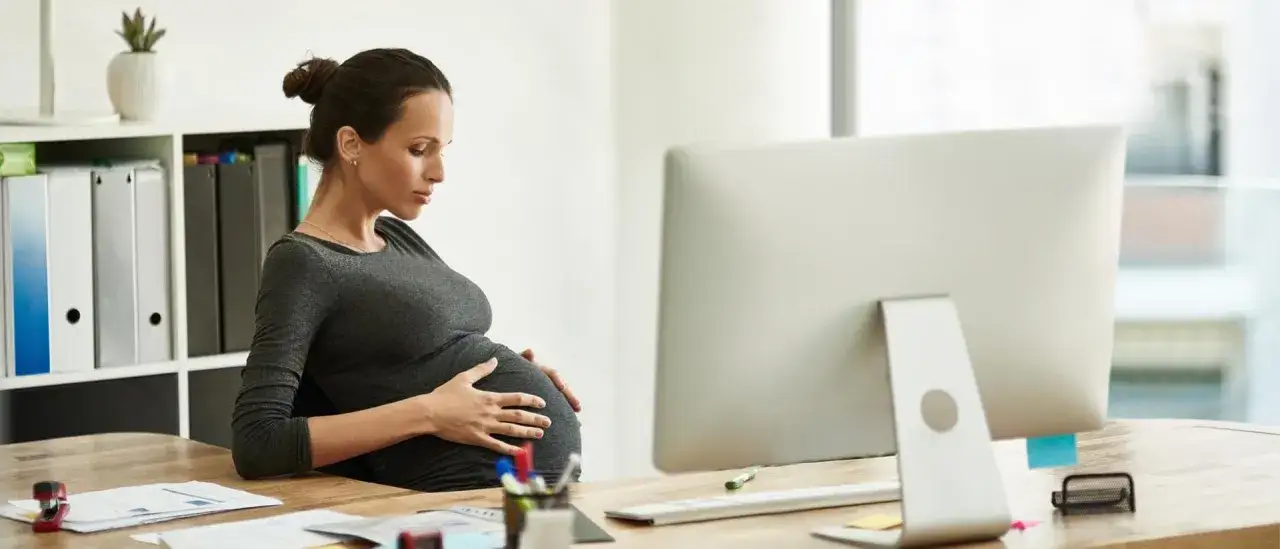 Gravid kvinna vid skrivbordet, med h&auml;nderna p&aring; magen, verkar uppleva gravid stress p&aring; jobb.