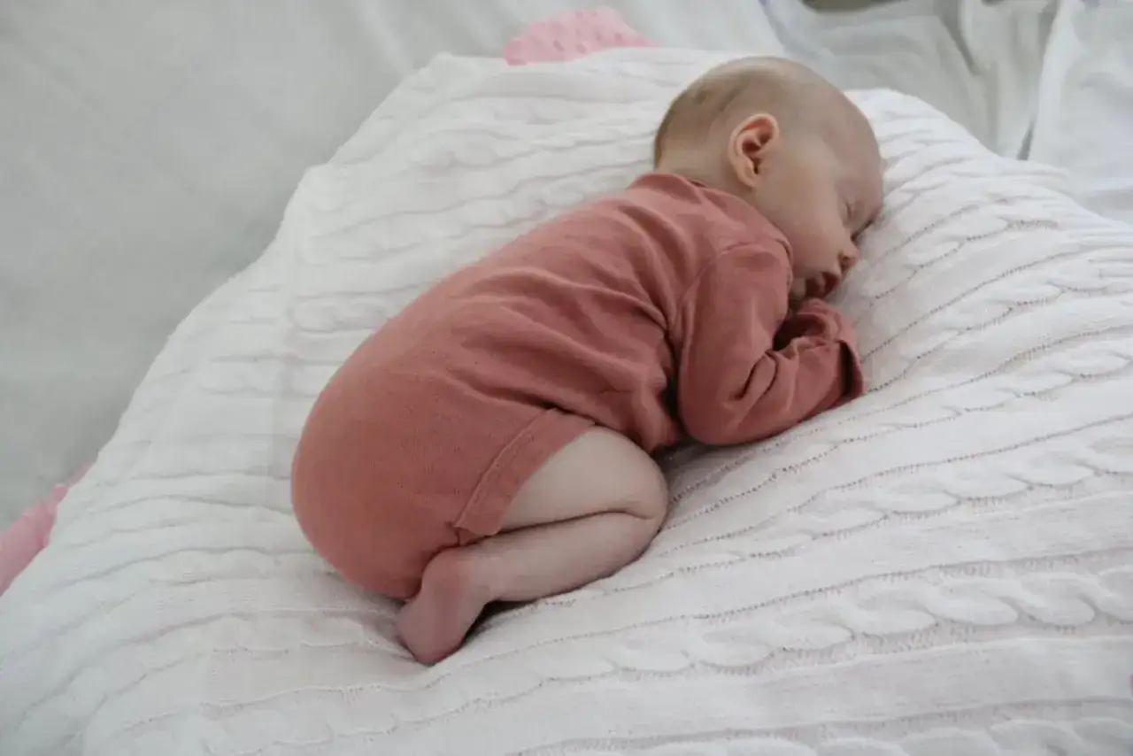 Sover s&ouml;tt p&aring; en vit stickad filt, en liten bebis i rosa body. Hoppas bebisen inte hosta.